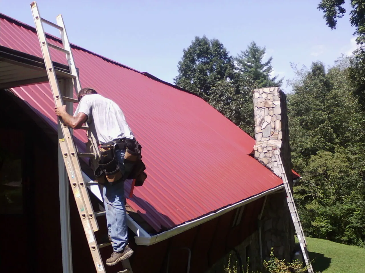 Roofer on a ladder beside a red metal roof and stone chimney — Soffit & Fascia Repair by Roofing Brothers in Monmouth Beach